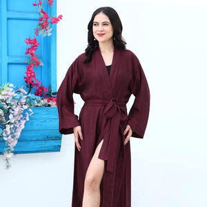 May include: A burgundy-colored robe with a matching belt, featuring long sleeves and a thigh-high slit. The robe is made of a lightweight fabric. The model is wearing beige sandals.