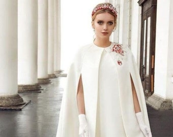 May include: A woman in a white dress and cape with a floral headpiece. The cape has arm openings and a decorative floral accent. She wears white gloves and heels. The background features white columns and a dark floor.