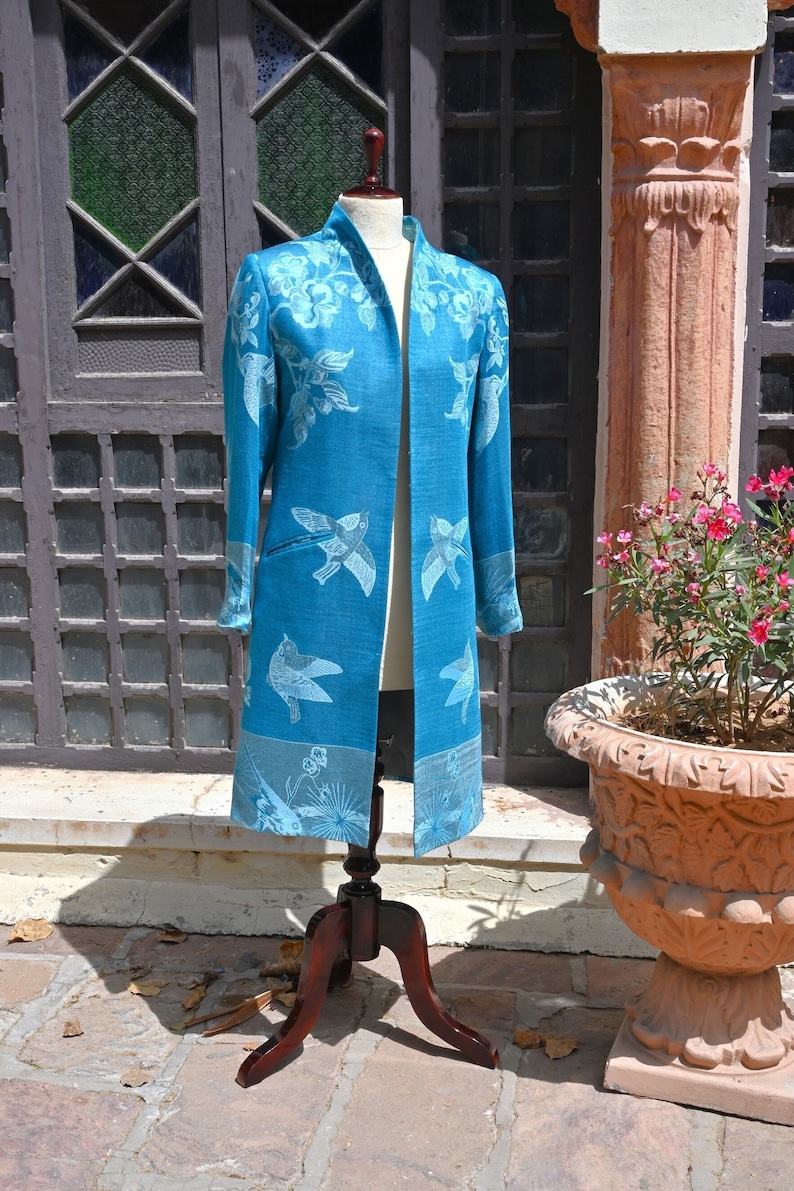 May include: A long, open-front teal coat with a white bird and floral pattern. The coat is displayed on a wooden dress form. The coat has long sleeves and a notched collar. The background includes a door and a decorative flower pot.