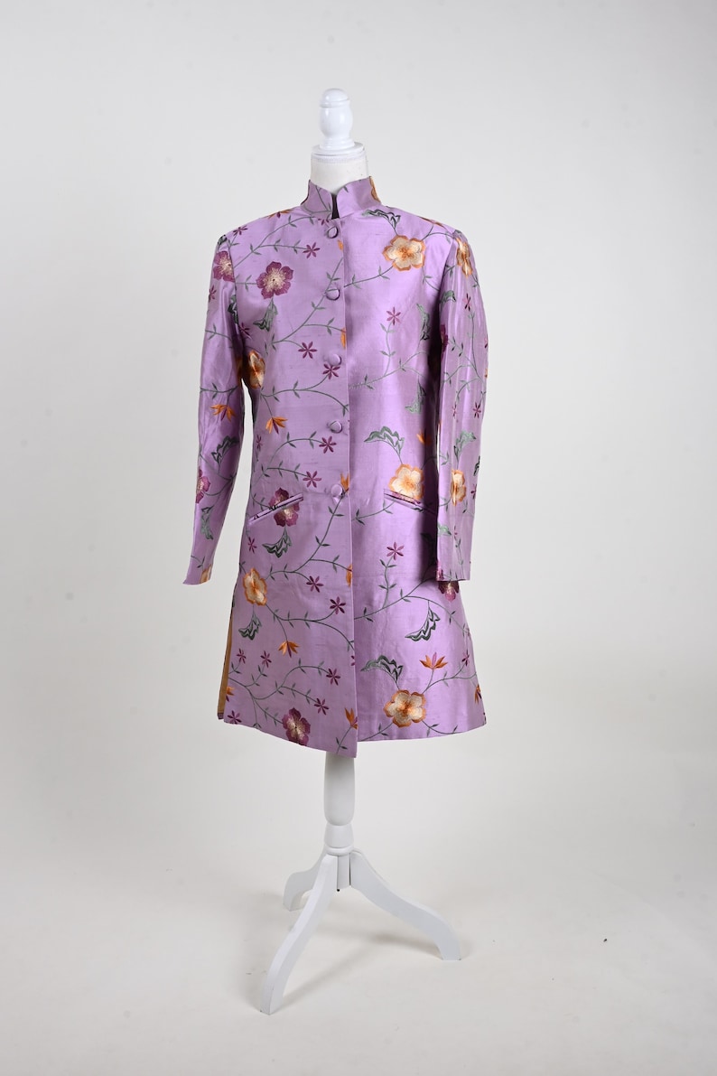 May include: A lavender coat with a mandarin collar and floral embroidery. The coat is displayed on a white mannequin. The embroidery features orange, purple, and green flowers and vines. The coat has long sleeves and a button-up front.