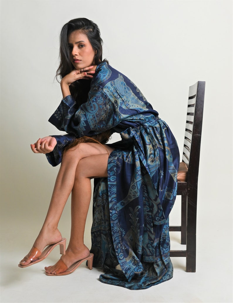 Puede incluir: Una bata larga y fluida de color azul con un estampado de cachemira en tonos azules y beige. La bata se lleva con sandalias de tac&oacute;n color nude. La modelo est&aacute; sentada en una silla de madera oscura sobre un fondo blanco.