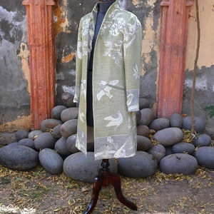 May include: A long, open-front coat in a pale green shade, decorated with white bird and floral designs. The coat is displayed on a wooden mannequin, highlighting its flowing shape. The coat has a contrasting grey border. The background features large stones and a weathered wall.