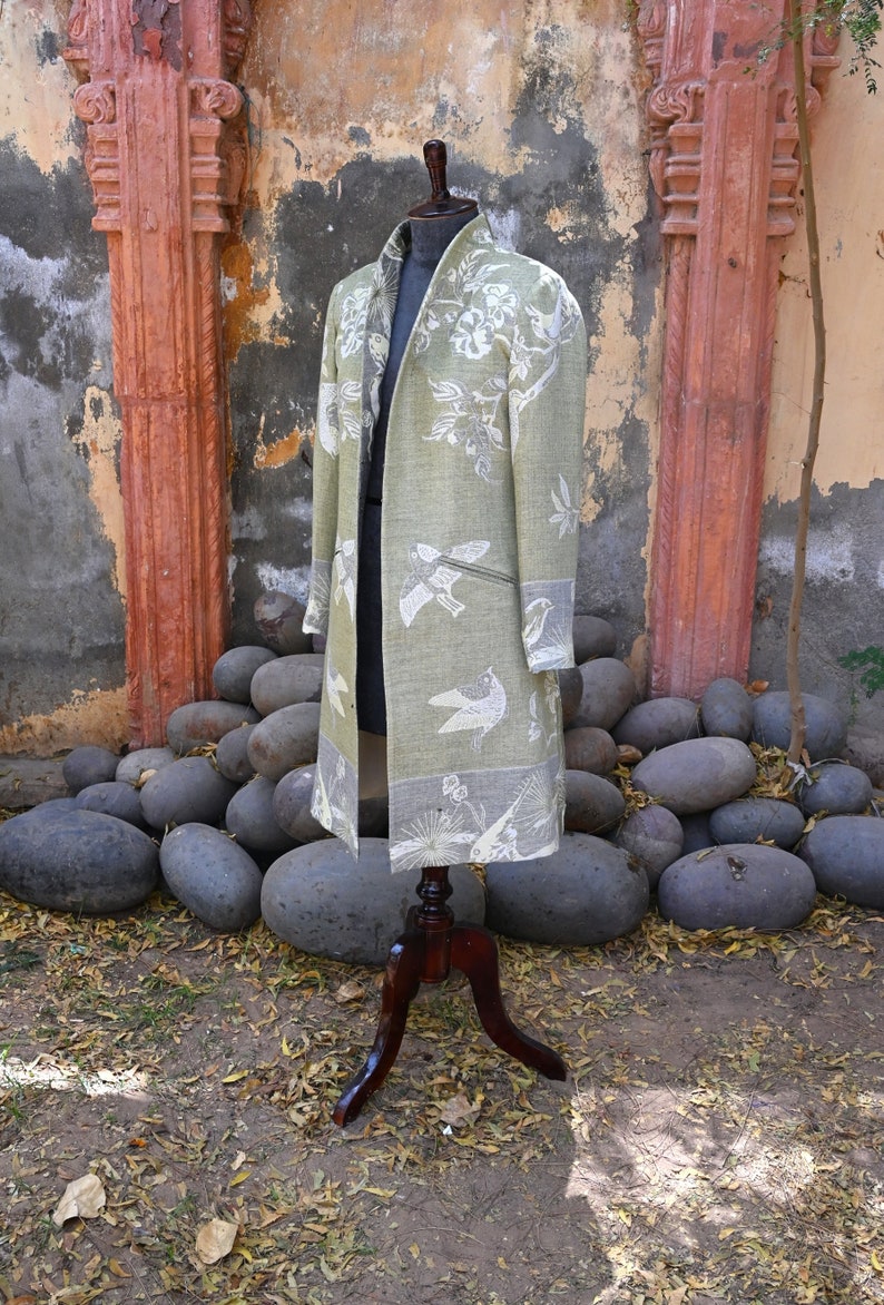 May include: A long, open-front coat in a pale green shade, decorated with white bird and floral designs. The coat is displayed on a wooden mannequin, highlighting its flowing shape. The coat has a contrasting grey border. The background features large stones and a weathered wall.