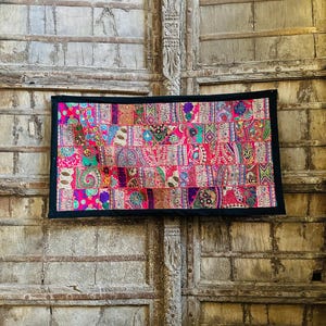 May include: A rectangular patchwork textile with a black border. The textile features a vibrant mix of colors, including pink, purple, green, and orange, arranged in a geometric pattern. The background is a weathered wooden door.