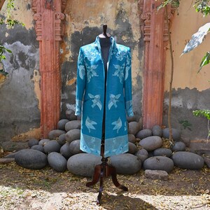 May include: A teal long coat with a floral and bird pattern, displayed on a wooden mannequin. The coat has a collar and pockets, and the bird design is repeated along the hem. The background features a stone wall and large grey rocks.