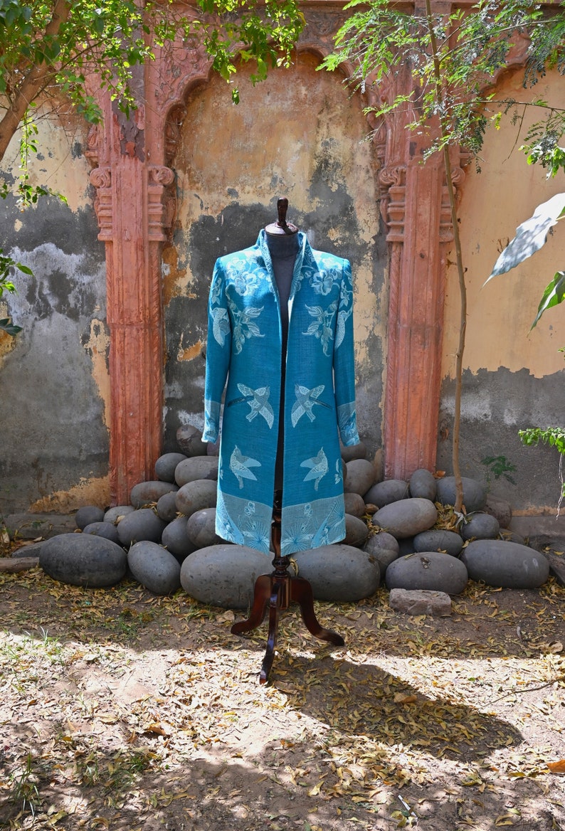 May include: A teal long coat with a floral and bird pattern, displayed on a wooden mannequin. The coat has a collar and pockets, and the bird design is repeated along the hem. The background features a stone wall and large grey rocks.
