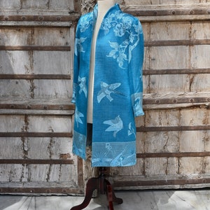 May include: A long, open-front blue jacket with a bird and floral pattern. The jacket is displayed on a mannequin with a dark wooden stand. The background features a weathered wooden door.