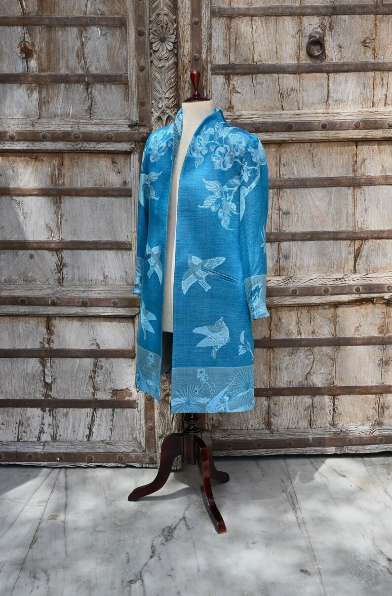 May include: A long, open-front blue jacket with a bird and floral pattern. The jacket is displayed on a mannequin with a dark wooden stand. The background features a weathered wooden door.