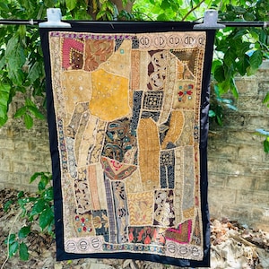 May include: A patchwork textile wall hanging with a black border. The design features various fabrics in shades of gold, brown, and beige, with accents of pink and red. The textile is rectangular and appears to be hanging outdoors.