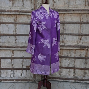 May include: A long, open-front purple jacket with a floral and bird pattern. The jacket is displayed on a mannequin with a wooden stand. The background is a weathered wooden door.