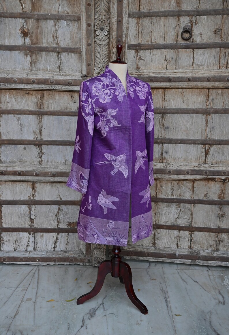 May include: A long, open-front purple jacket with a floral and bird pattern. The jacket is displayed on a mannequin with a wooden stand. The background is a weathered wooden door.