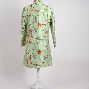 May include: A light green tunic with long sleeves, displayed on a white mannequin. The tunic is adorned with an embroidered floral pattern in shades of orange and brown. The garment has a mandarin collar and a straight hemline.