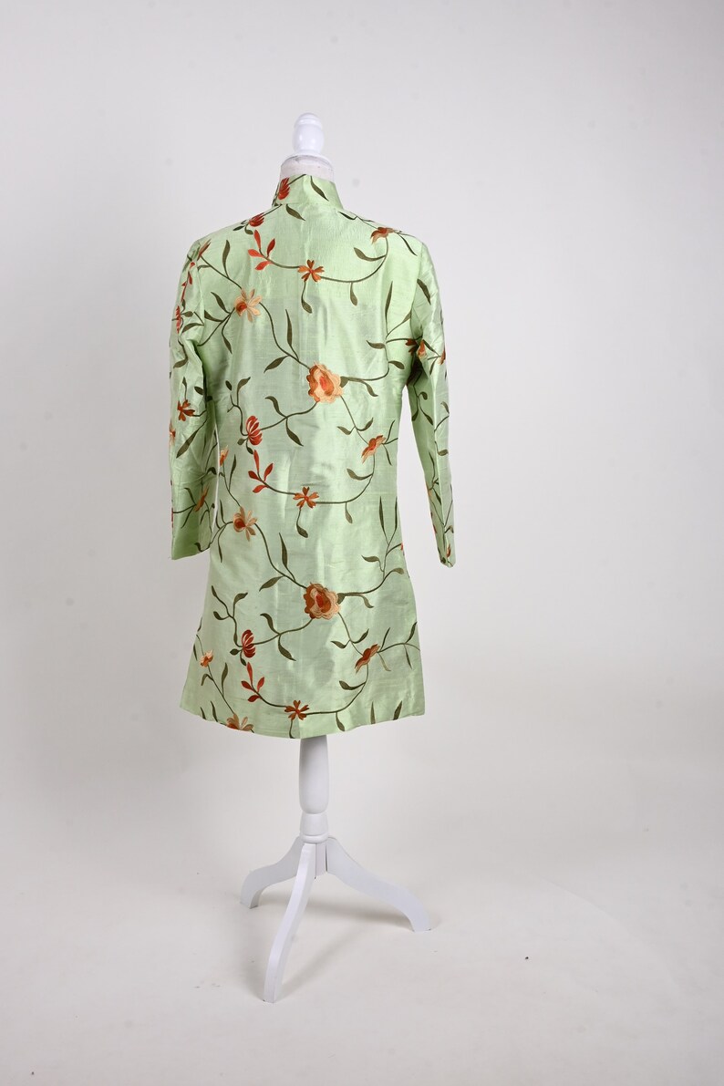 May include: A light green tunic with long sleeves, displayed on a white mannequin. The tunic is adorned with an embroidered floral pattern in shades of orange and brown. The garment has a mandarin collar and a straight hemline.