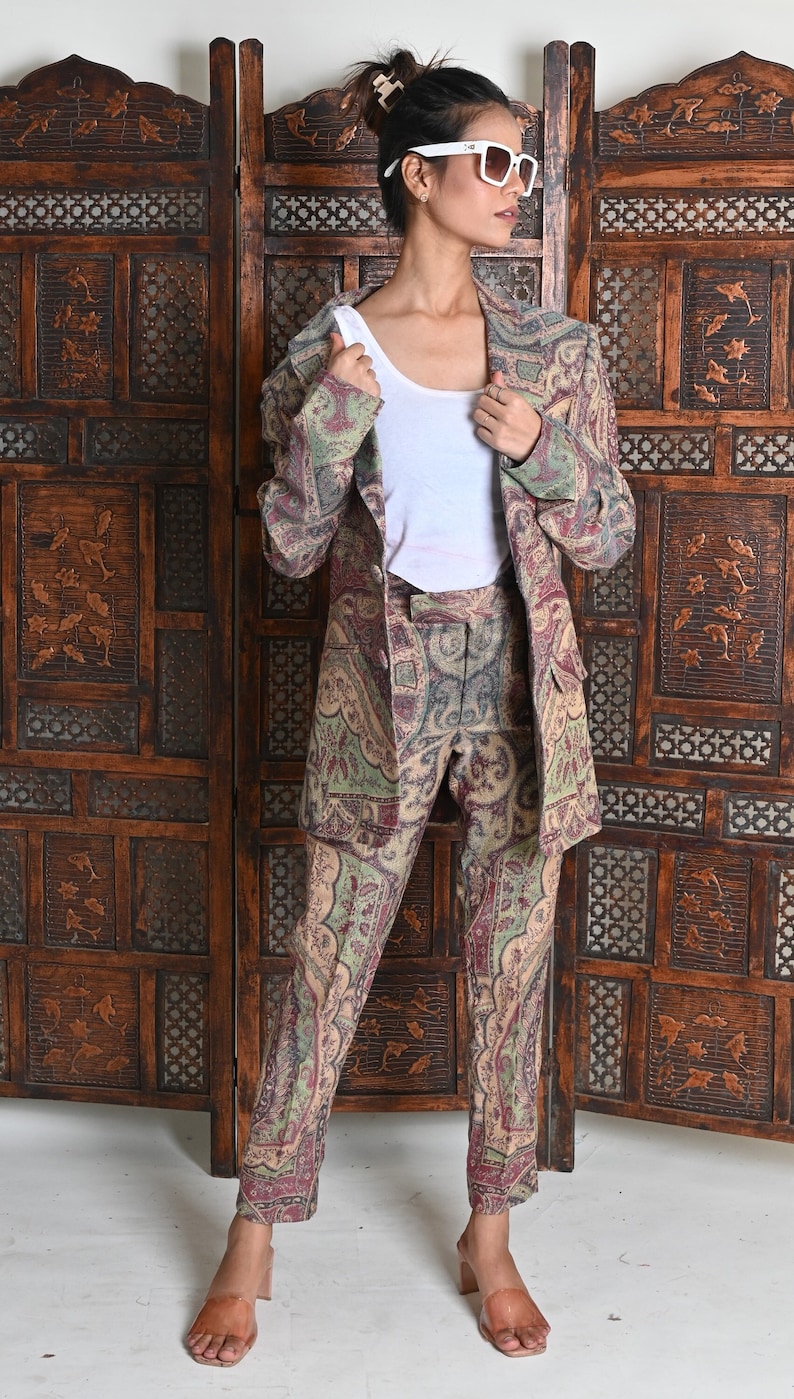 May include: A paisley-patterned suit in shades of green, burgundy, and beige. The suit includes a blazer and matching trousers. The person is wearing a white top and white sunglasses. The background features a wooden room divider.