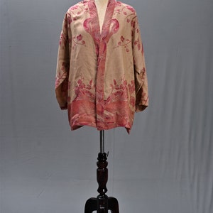 May include: A beige kimono-style jacket with a floral pattern in shades of pink and red. The jacket is displayed on a wooden mannequin stand. The floral design is concentrated along the edges and front opening.