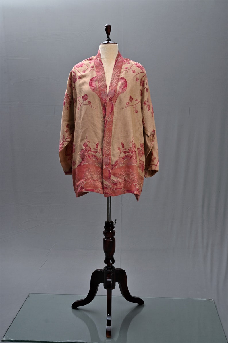May include: A beige kimono-style jacket with a floral pattern in shades of pink and red. The jacket is displayed on a wooden mannequin stand. The floral design is concentrated along the edges and front opening.