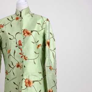 May include: A light green, long-sleeved jacket with a mandarin collar, adorned with embroidered floral patterns in shades of orange and brown. The jacket is displayed on a mannequin, showcasing its design and fit. The garment has two front pockets.