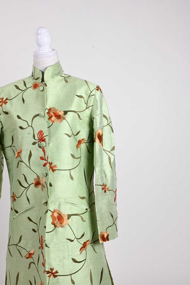 May include: A light green, long-sleeved jacket with a mandarin collar, adorned with embroidered floral patterns in shades of orange and brown. The jacket is displayed on a mannequin, showcasing its design and fit. The garment has two front pockets.