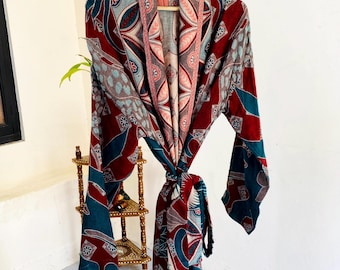 May include: A patterned robe in shades of red, teal, and gray hangs on a wooden hanger. The robe features a repeating geometric design and is tied at the waist. The robe is made of a lightweight fabric.