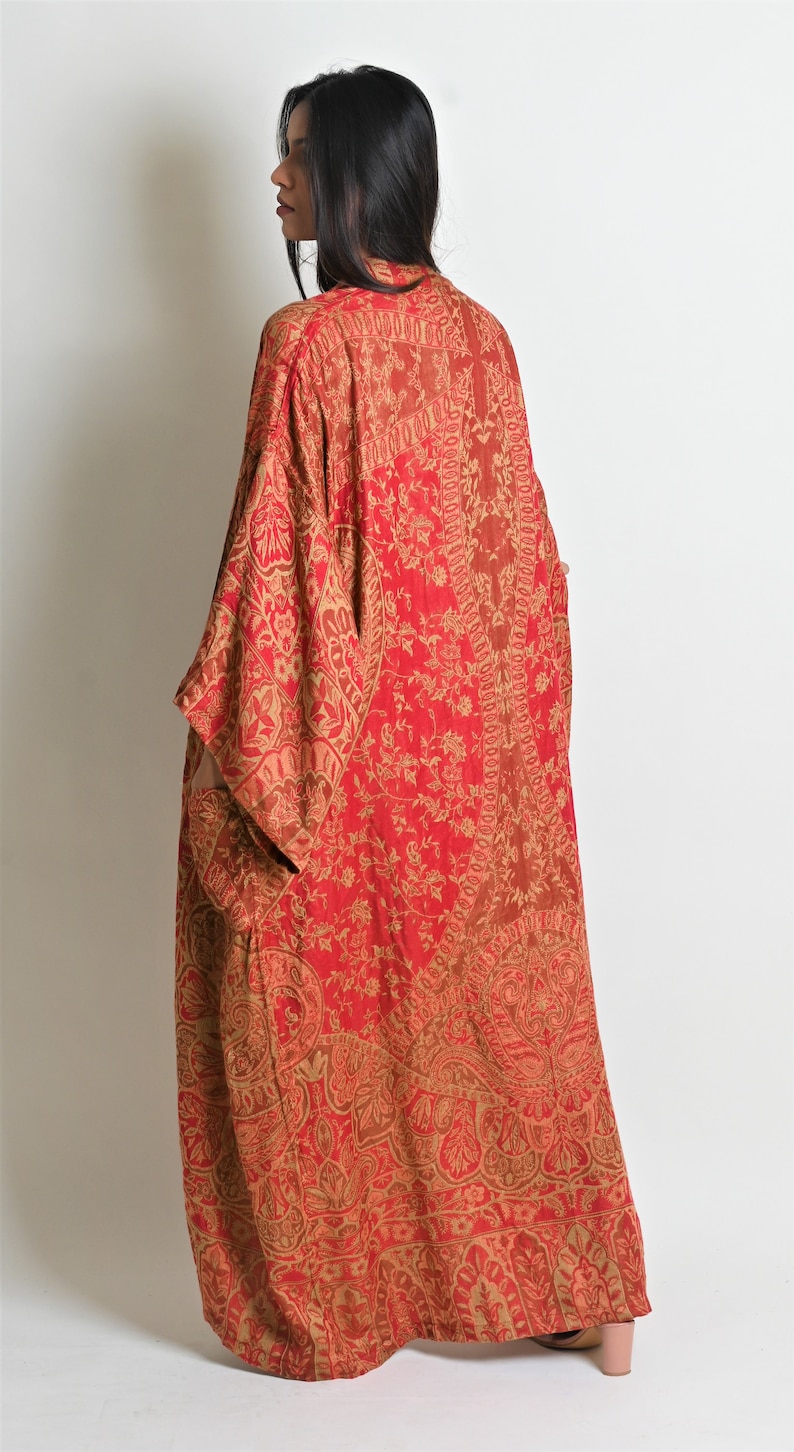 May include: A long, flowing red and gold patterned kimono-style robe. The robe features an intricate paisley and floral design. The garment is made of a lightweight fabric and has wide sleeves. The model is wearing the robe.