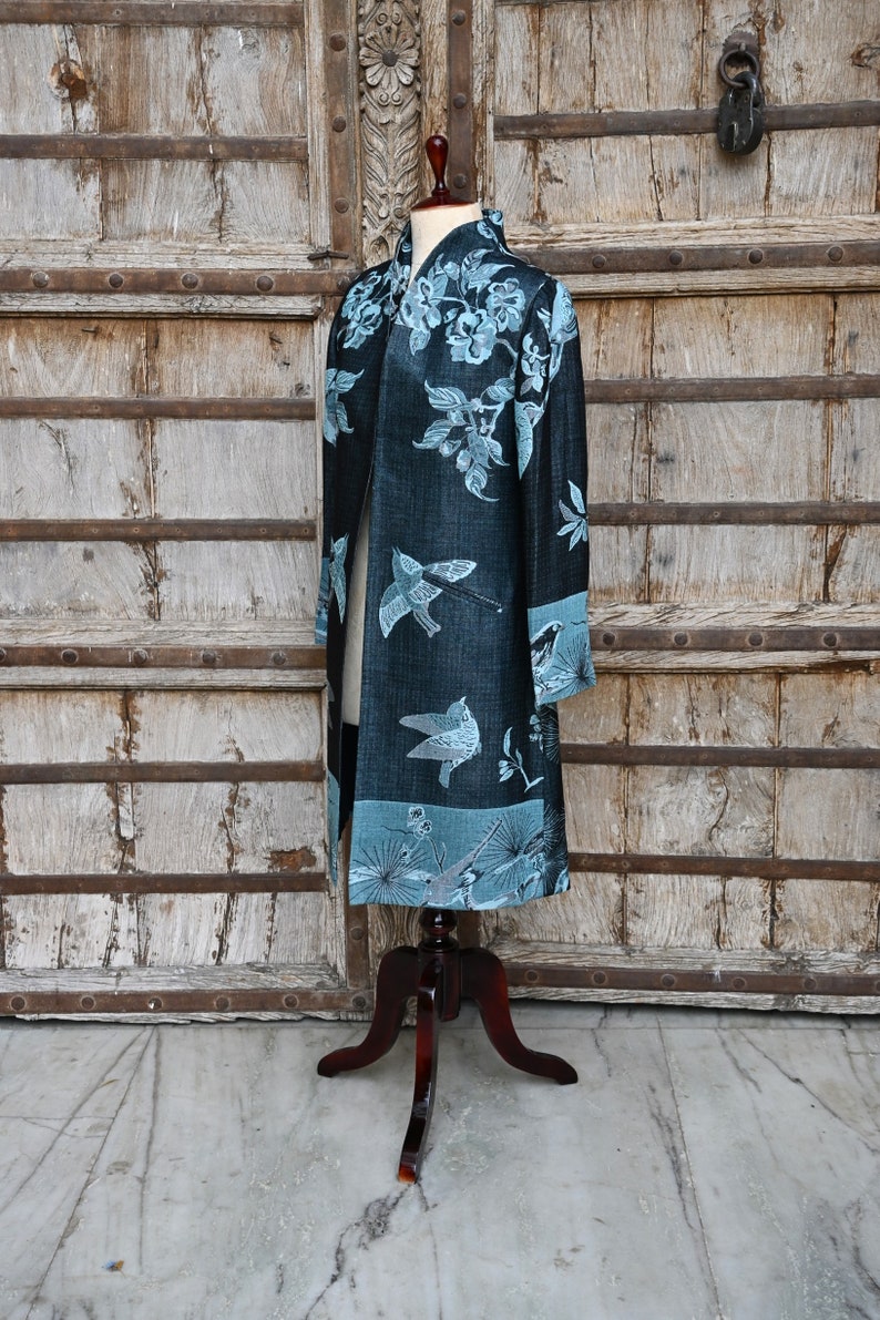 May include: A long, dark blue kimono-style robe with a floral and bird pattern in light blue. The robe is displayed on a wooden mannequin, showcasing its design and length. The background features a rustic wooden door.