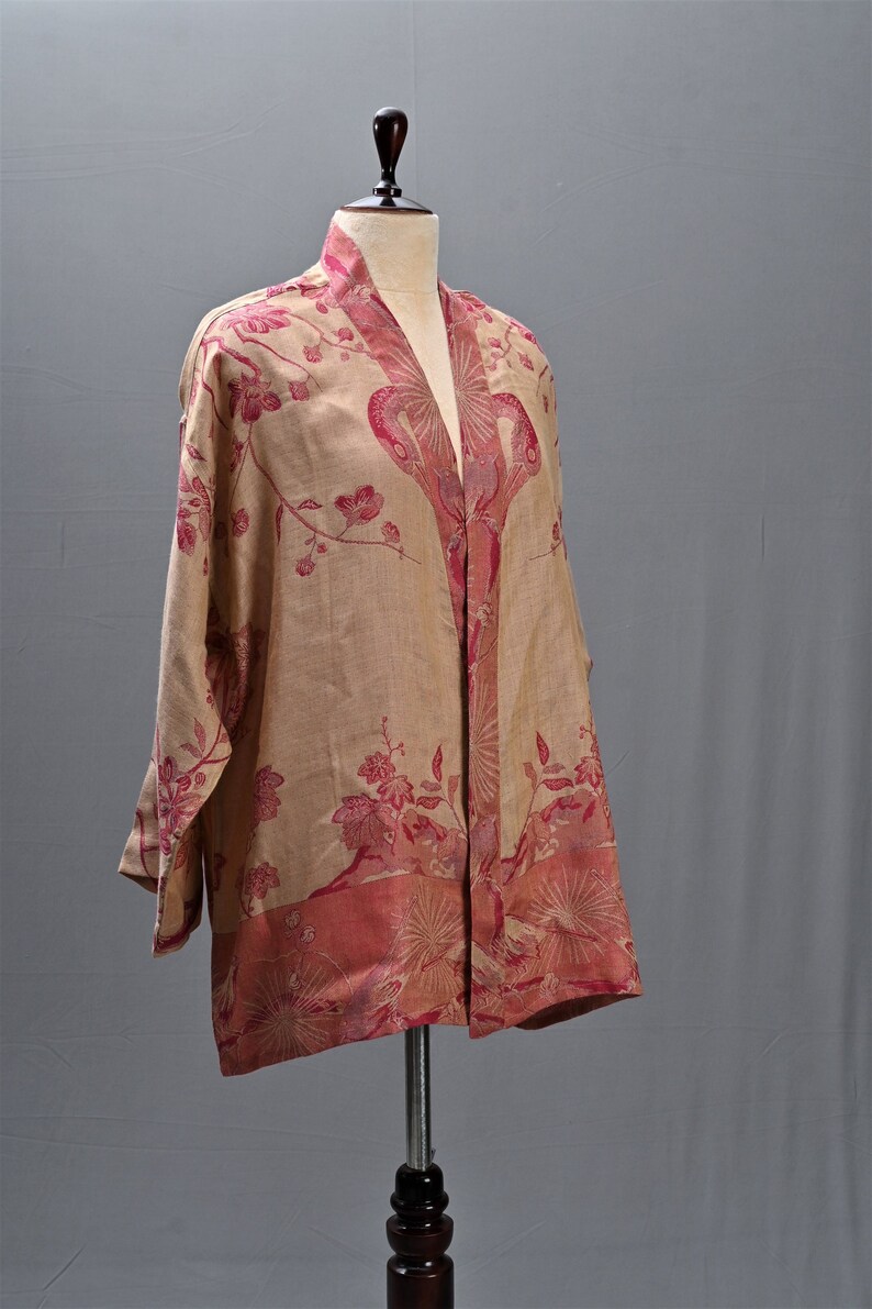 May include: A beige kimono-style jacket with a floral pattern in shades of pink and red. The jacket is displayed on a mannequin, showcasing its open front and long sleeves. The floral design is concentrated on the borders and along the front.