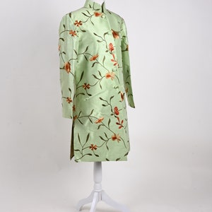 May include: A light green, long-sleeved jacket with a mandarin collar, displayed on a white mannequin. The jacket is adorned with a floral pattern in shades of orange and brown. The garment has a straight cut and side slits.