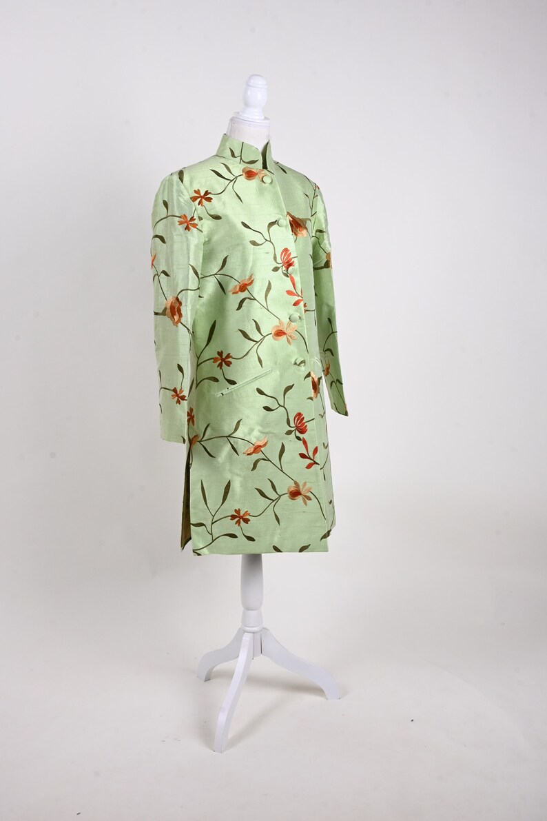 May include: A light green, long-sleeved jacket with a mandarin collar, displayed on a white mannequin. The jacket is adorned with a floral pattern in shades of orange and brown. The garment has a straight cut and side slits.