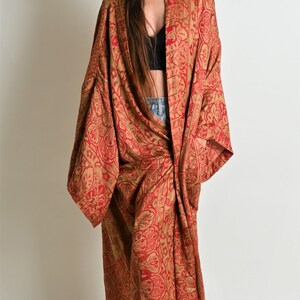 May include: A long, flowing red and gold patterned kimono-style robe. The robe features an intricate paisley design and is worn open, revealing a black top and denim shorts. The garment drapes to the floor.