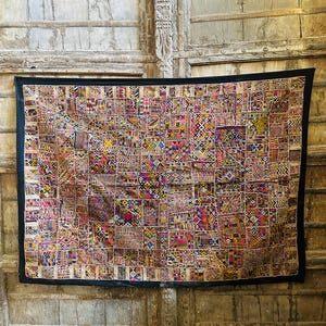 Puede incluir: Un textil rectangular multicolor con un borde negro. Está compuesto por numerosos parches cuadrados pequeños, cada uno con un patrón geométrico único en tonos de rosa, amarillo, azul y marrón. El fondo es una puerta de madera envejecida.