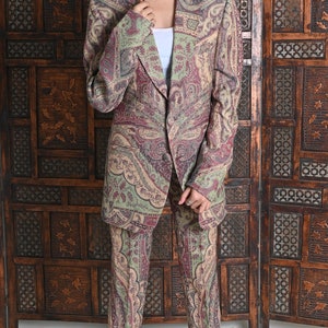 May include: A paisley-patterned suit in shades of burgundy, green, and beige. The suit includes a blazer and matching trousers. The person is wearing white sunglasses and clear heeled sandals. The background features a wooden screen.