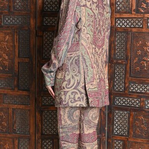 May include: A paisley-patterned suit in shades of beige, burgundy, and green. The suit includes a blazer and matching pants. The person is wearing sunglasses and heels. The background features a wooden screen.