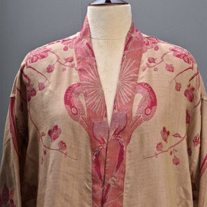 May include: A beige kimono-style robe with a floral and bird pattern in shades of pink. The robe is displayed on a mannequin, showcasing the intricate design and flowing fabric. The robe's collar and edges are accented with a darker pink trim.