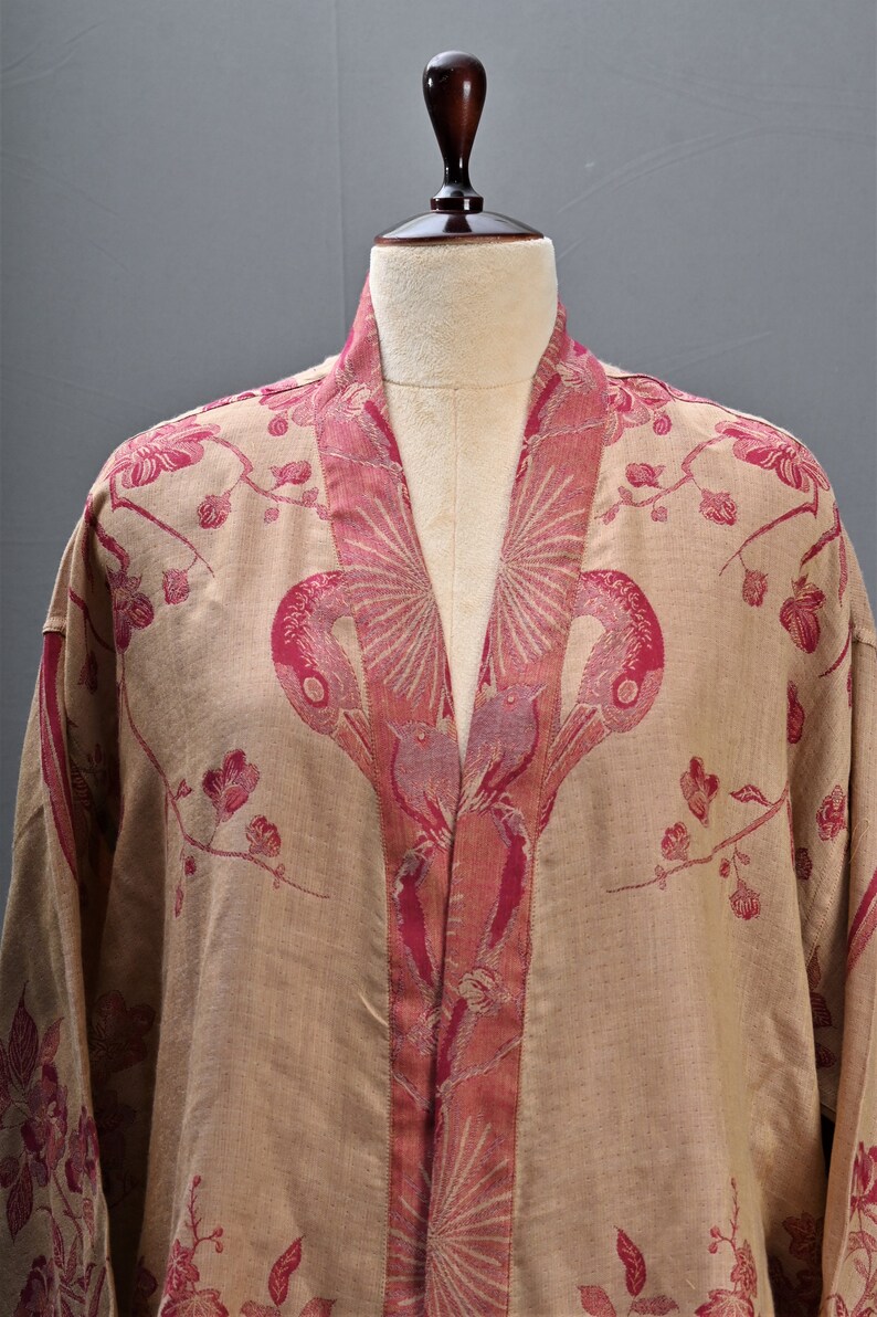May include: A beige kimono-style robe with a floral and bird pattern in shades of pink. The robe is displayed on a mannequin, showcasing the intricate design and flowing fabric. The robe's collar and edges are accented with a darker pink trim.