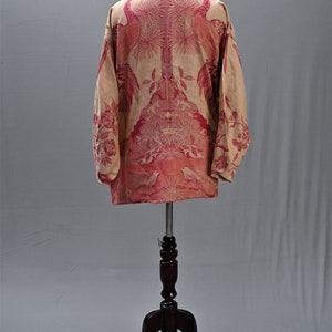 May include: A beige long-sleeved shirt with a pink floral and bird pattern. The shirt is displayed on a wooden mannequin. The design features birds, flowers, and other botanical elements.