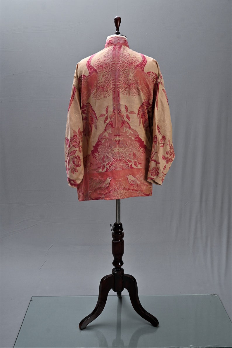 May include: A beige long-sleeved shirt with a pink floral and bird pattern. The shirt is displayed on a wooden mannequin. The design features birds, flowers, and other botanical elements.