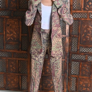 May include: A paisley-patterned suit in shades of olive green, burgundy, and beige. The suit consists of a blazer and matching trousers. The person in the image is wearing a white tank top and white sunglasses. The background features a wooden screen.