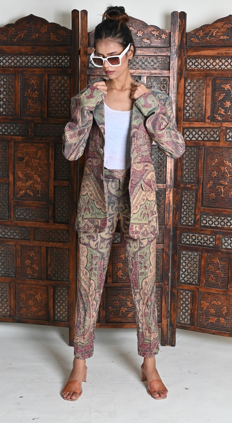 May include: A paisley-patterned suit in shades of olive green, burgundy, and beige. The suit consists of a blazer and matching trousers. The person in the image is wearing a white tank top and white sunglasses. The background features a wooden screen.