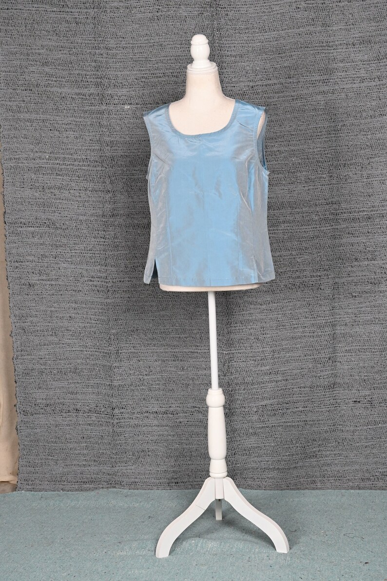 Pu&ograve; includere: Un top senza maniche azzurro chiaro con scollo tondo &egrave; esposto su un manichino bianco. Il top ha una leggera lucentezza e uno spacco laterale. Il manichino &egrave; su un supporto bianco con base a treppiede, su uno sfondo grigio strutturato.