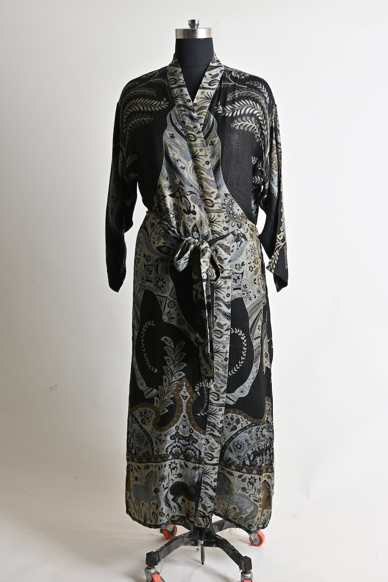 Puede incluir: Una bata larga negra estampada con un dise&ntilde;o envolvente, exhibida en un maniqu&iacute;. La bata presenta un patr&oacute;n detallado en tonos grises, beige y verde oliva. La tela est&aacute; atada a la cintura con una faja a juego. La bata es larga hasta el suelo.