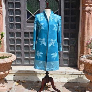 May include: A teal duster coat with a floral and bird pattern, displayed on a wooden mannequin. The coat has long sleeves, a collarless design, and a knee-length hem. The background includes a wooden door and a stone patio.