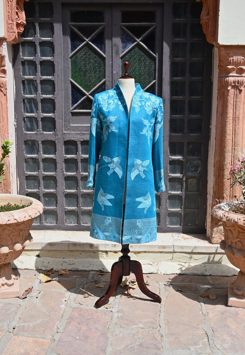 May include: A teal duster coat with a floral and bird pattern, displayed on a wooden mannequin. The coat has long sleeves, a collarless design, and a knee-length hem. The background includes a wooden door and a stone patio.