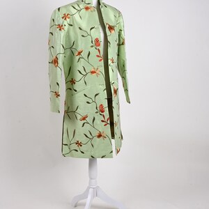 May include: A light green, long-sleeved jacket with a mandarin collar and floral embroidery. The jacket is displayed on a white mannequin. The embroidery features brown stems and orange flowers. The jacket has a straight cut and side slits.
