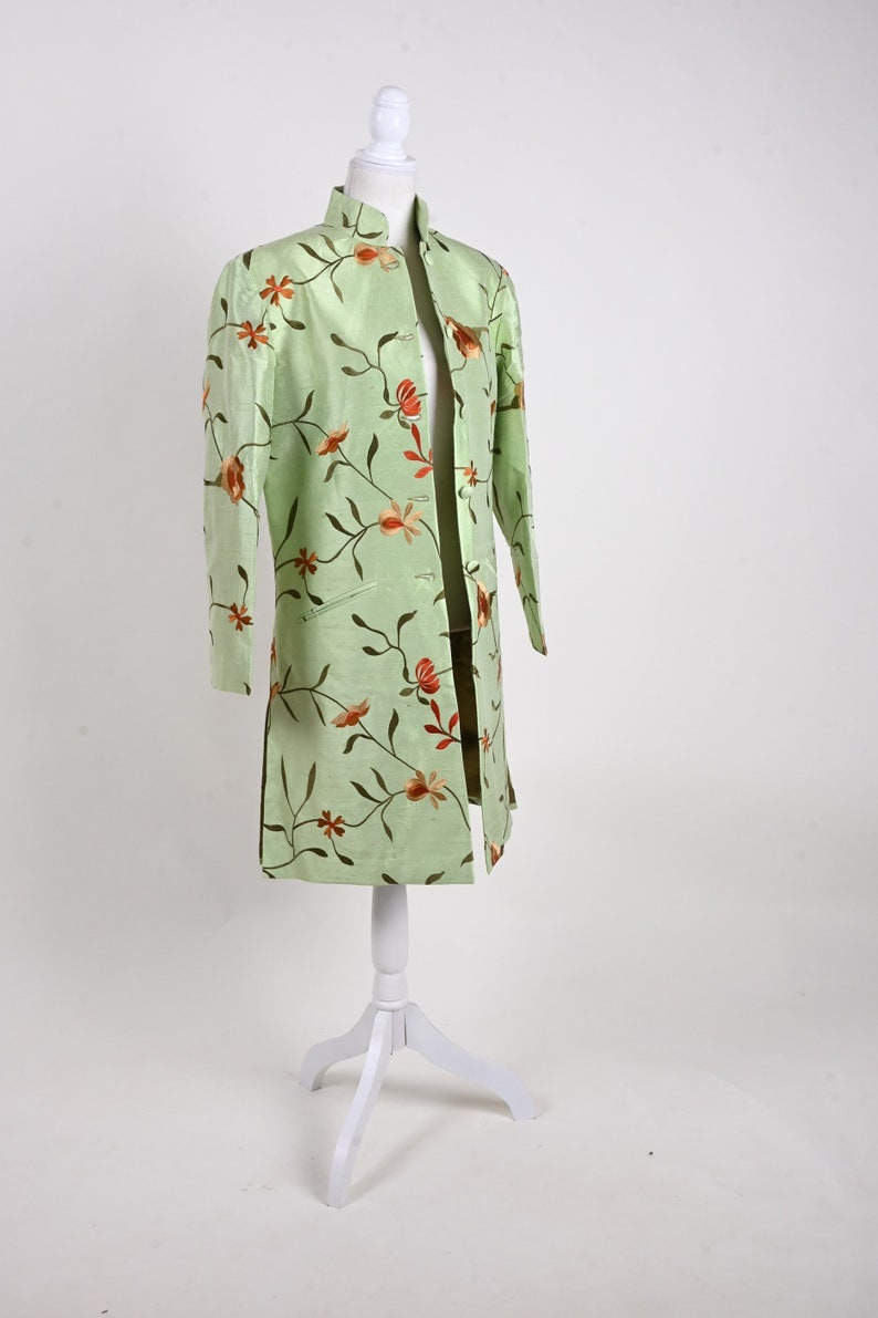 May include: A light green, long-sleeved jacket with a mandarin collar and floral embroidery. The jacket is displayed on a white mannequin. The embroidery features brown stems and orange flowers. The jacket has a straight cut and side slits.