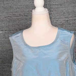 Pu&ograve; includere: Un top senza maniche azzurro chiaro con scollatura rotonda. Il tessuto ha una lucentezza sottile e l'indumento &egrave; esposto su un manichino. Lo sfondo &egrave; un tessuto grigio strutturato.