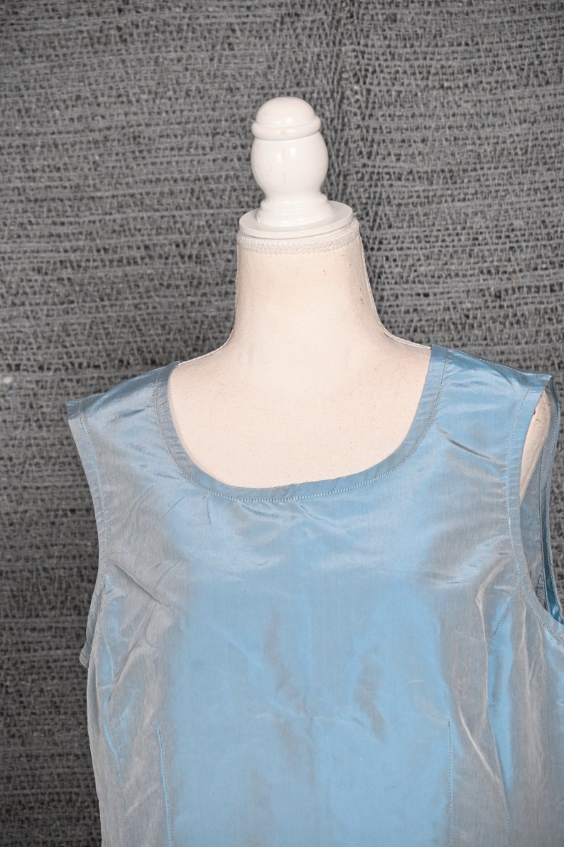 Pu&ograve; includere: Un top senza maniche azzurro chiaro con scollatura rotonda. Il tessuto ha una lucentezza sottile e l'indumento &egrave; esposto su un manichino. Lo sfondo &egrave; un tessuto grigio strutturato.