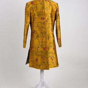 May include: Mustard yellow long coat with a mandarin collar and floral embroidery. The coat features a button-down front and a split hem. The floral design includes red flowers and green stems. The coat is displayed on a white mannequin.