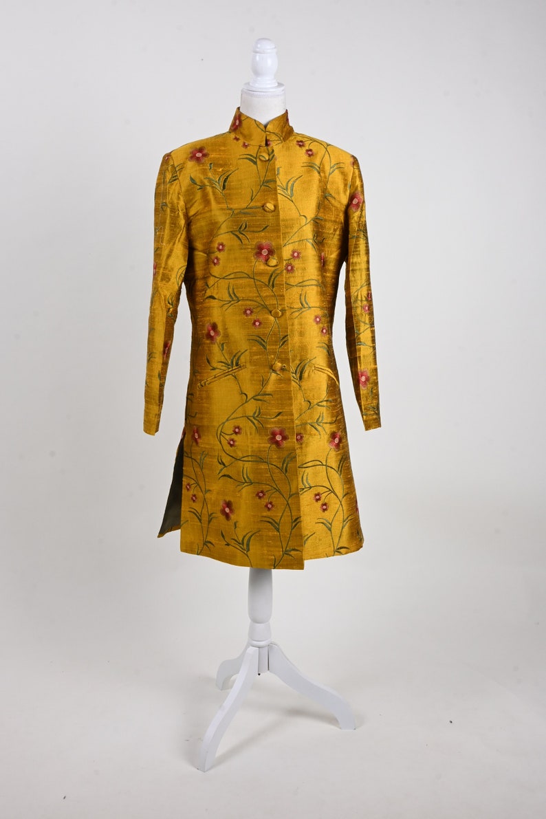 May include: Mustard yellow long coat with a mandarin collar and floral embroidery. The coat features a button-down front and a split hem. The floral design includes red flowers and green stems. The coat is displayed on a white mannequin.