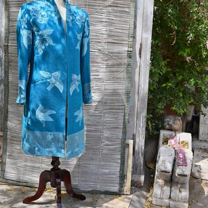 May include: A teal long coat with a floral and bird design is displayed on a wooden mannequin. The coat features a mandarin collar and long sleeves. The background includes a bamboo screen and a wooden door.