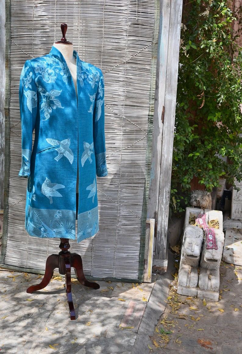 May include: A teal long coat with a floral and bird design is displayed on a wooden mannequin. The coat features a mandarin collar and long sleeves. The background includes a bamboo screen and a wooden door.